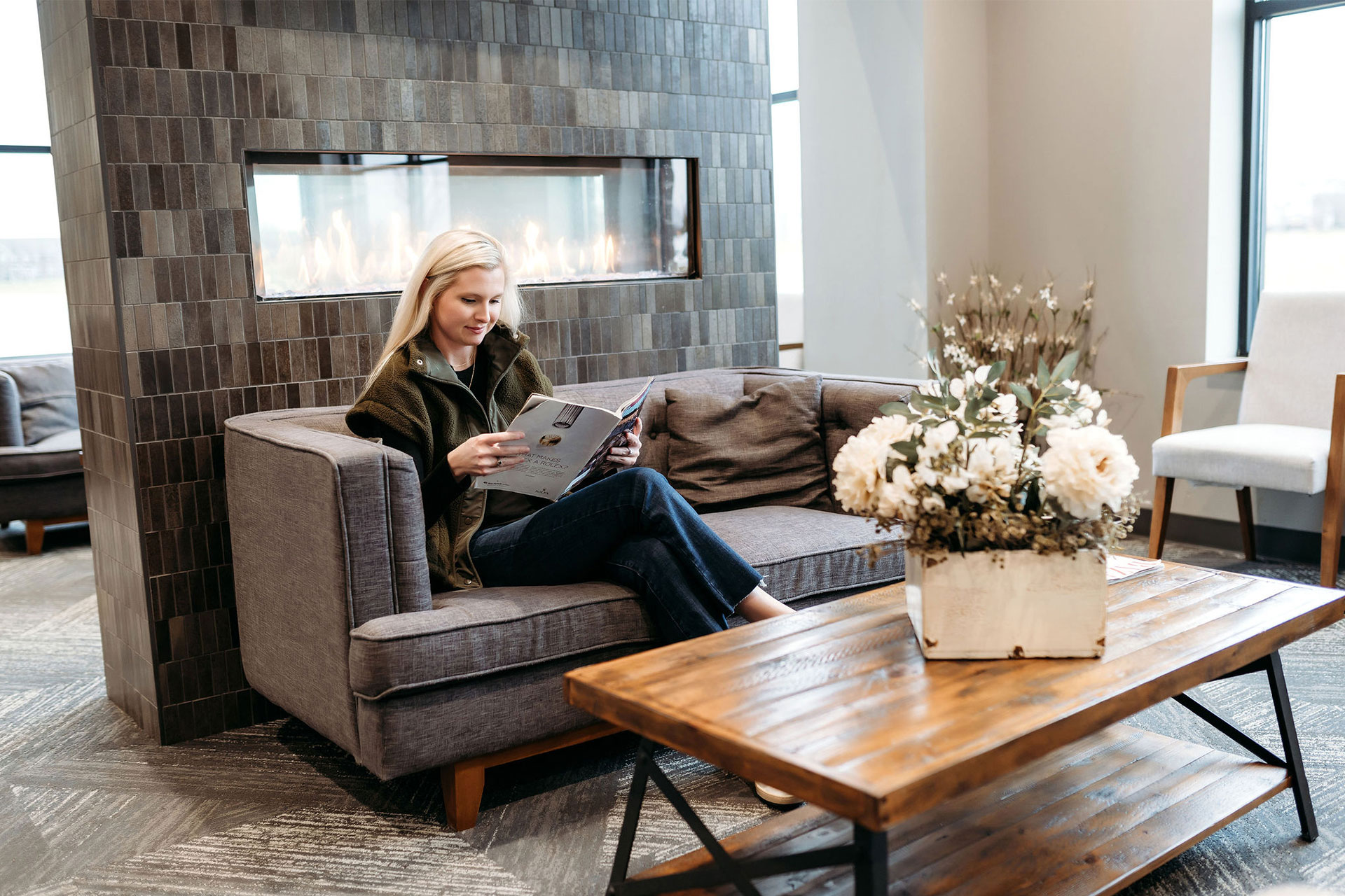 vds-office-2 Woman reading in a cozy Village Dental lounge with a modern fireplace and floral décor.